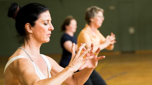 Drei Frauen führen eine Fitnessübung in einem Saal mit Holzfußboden und einer Sporttorwand im Hintergrund aus.
