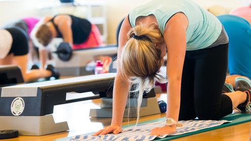 Frau in Fitnesskleidung übt auf ihrer Matte in einem Gruppenfitnesskurs mit weiteren Teilnehmern im Hintergrund.