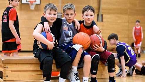 Drei Kinder posieren auf einer Bank in einer Sporthalle, jeder hält einen Basketball. Im Hintergrund weitere Spieler.