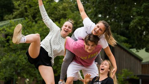 Vier junge Frauen posieren fröhlich auf einer sandigen Fläche, eine sitzt im Spagat, während die anderen sie umarmen.