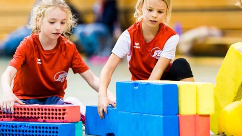 Drei Mädchen im roten Shirt bauen mit bunten Schaumstoffbausteinen in einer Turnhalle.