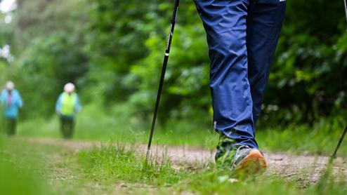 Nahaufnahme eines wandernden Fußes mit Trekkingstock auf einem schmalen Weg, im Hintergrund zwei weitere Personen.