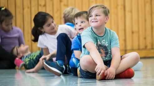 Gruppe von Kindern sitzt in einer Turnhalle, einige lachen, während ein Junge mit roten Schuhen lächelt.