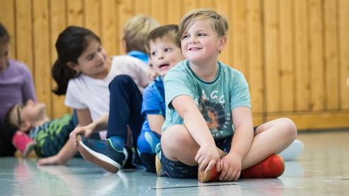 Gruppe von Kindern sitzt in einer Turnhalle, einige lachen, während ein Junge mit roten Schuhen lächelt.