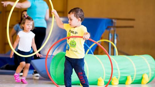 Zwei Kinder spielen in einer Turnhalle mit hula-hoops und einem grünen Tunnel. Ein Kind hält zwei Reifen in der Hand.