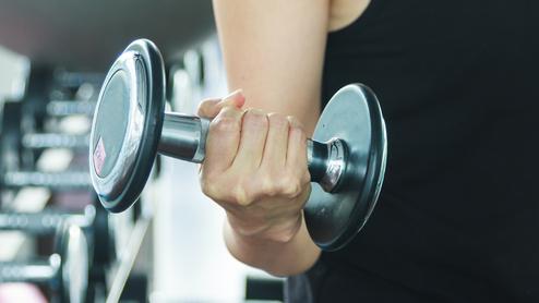 Nahaufnahme einer Hand, die eine Kurzhantel im Fitnessstudio hält, mit weiteren Hanteln im Hintergrund.