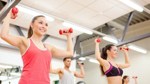 Vier Personen heben Gewichte in einem Fitnessstudio. Zwei Frauen tragen Sporttops, ein Mann hat ein T-Shirt an.