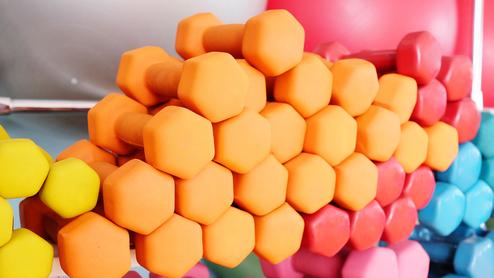 Bunte Hanteln in verschiedenen Farben und Größen, ordentlich auf einem Regal in einem Fitnessraum angeordnet.