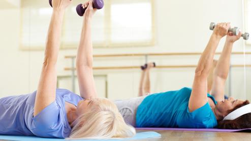 Zwei Frauen liegen auf Gymnastikmatten und heben Gewichte in einer Fitnessumgebung.