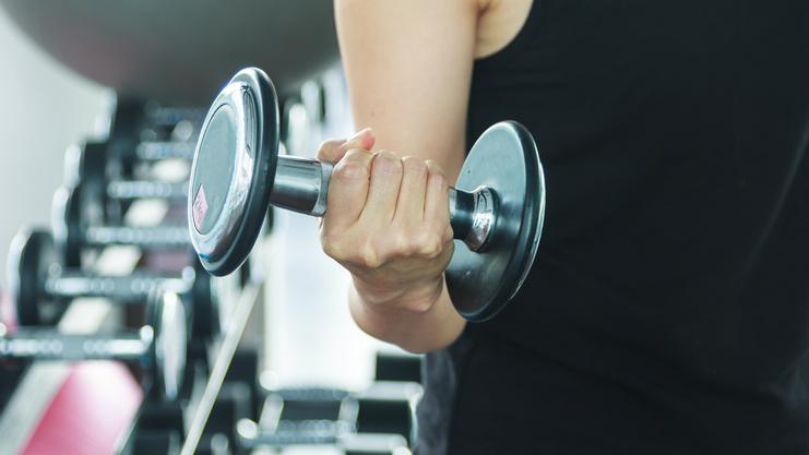 Nahaufnahme einer Hand, die eine Kurzhantel im Fitnessstudio hält, mit weiteren Hanteln im Hintergrund.