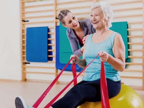 Frau mit grauen Haaren sitzt auf einem großen Gymnastikball, während eine Trainerin ihr mit engen Bändern hilft.