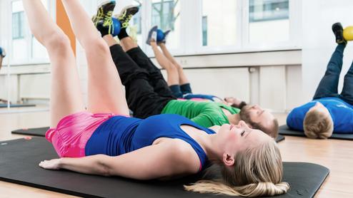 Gruppentraining: Frauen und Männer liegen auf Matten, heben Beine mit Medizinbällen in einem modernen Fitnessstudio.