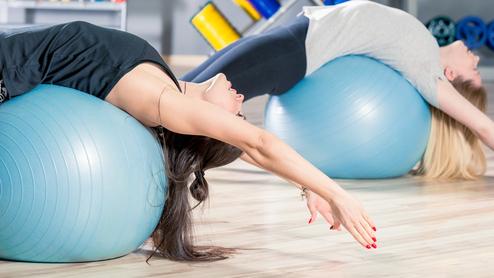 Zwei Frauen dehnen sich auf blauen Gymnastikbällen in einem Fitnessstudio mit Holzfußboden.
