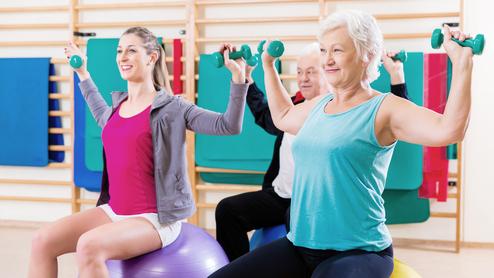 Drei Personen mit Hanteln sitzen auf großen Gymnastikbällen und trainieren in einem Fitnessraum.