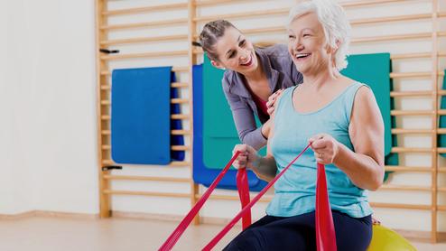 Frau mit grauen Haaren sitzt auf einem großen Gymnastikball, während eine Trainerin ihr mit engen Bändern hilft.