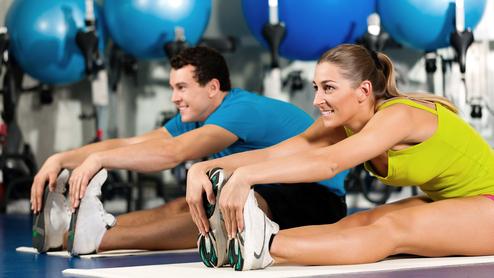 Zwei Personen dehnen auf Matten in einem Fitnessstudio, bunte Gymnastikbälle im Hintergrund.