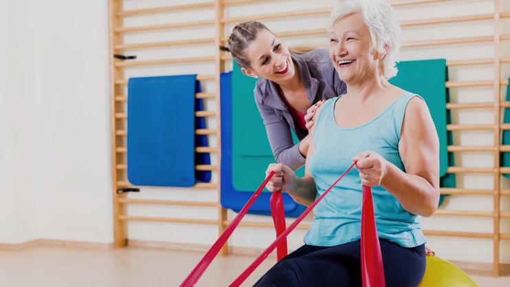 Frau mit grauen Haaren sitzt auf einem großen Gymnastikball, während eine Trainerin ihr mit engen Bändern hilft.