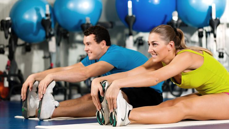 Zwei Personen dehnen auf Matten in einem Fitnessstudio, bunte Gymnastikbälle im Hintergrund.