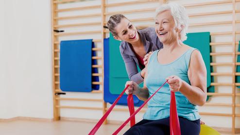 Frau mit grauen Haaren sitzt auf einem großen Gymnastikball, während eine Trainerin ihr mit engen Bändern hilft.