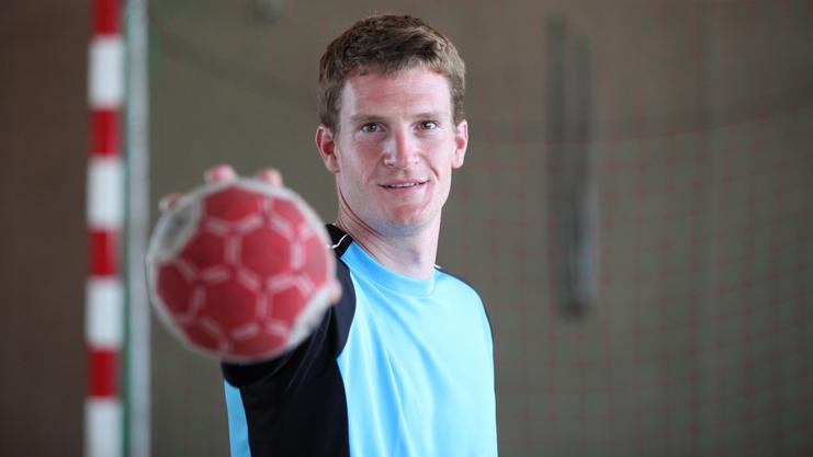 Sportler in blauem Trikot mit Handball in der Hand, lächelt in einer Sporthalle vor dem Tor.