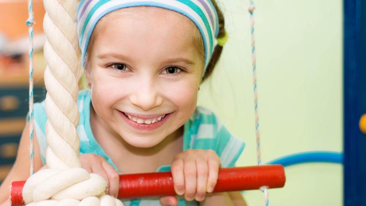 Lächelndes Mädchen mit blauer Kopfband und gestreifter Bluse hält sich an einem Klettergerüst fest.