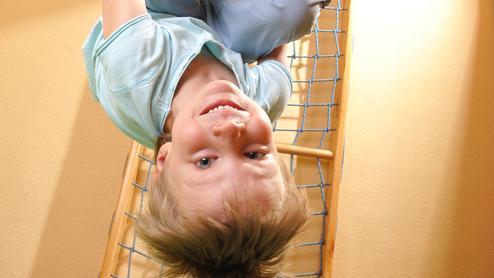 Kind hängt kopfüber an Turnringen, lächelt und trägt hellblaue Kleidung, vor runder Turngerüst-Wand mit Netz.