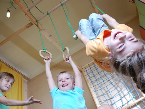 Drei Kinder spielen in einem Raum: Ein Junge hängt an Ringen, während zwei andere lachen und zuschauen.