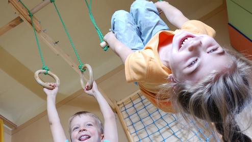 Drei Kinder spielen in einem Raum: Ein Junge hängt an Ringen, während zwei andere lachen und zuschauen.