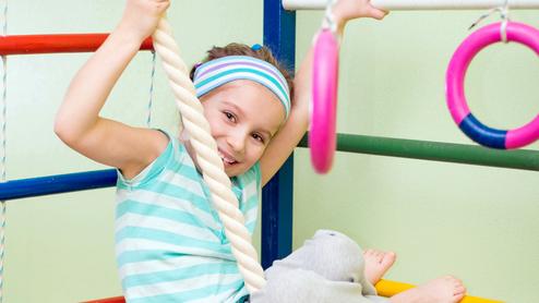Ein Mädchen hängt lachend an einem Kletterseil in einer Turnhalle, umgeben von bunten Turngeräten.