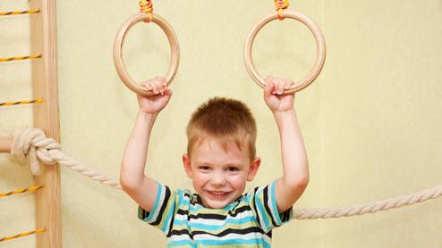 Junge mit einem gestreiften T-Shirt hält sich an zwei Holzringen in einem Spielraum fest und lächelt.