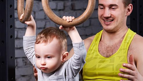Ein Erwachsener unterstützt ein Kind, das an Holzringen im Fitnessstudio hängt, beide lächeln.