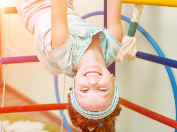 Mädchen hängt kopfüber an einem Spielgerüst und lächelt, während sie rosa Ringe in den Händen hält.