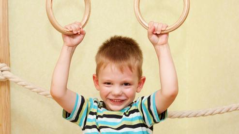 Junge mit einem gestreiften T-Shirt hält sich an zwei Holzringen in einem Spielraum fest und lächelt.