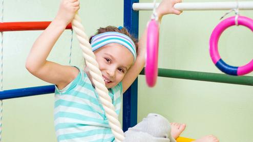 Ein Mädchen hängt lachend an einem Kletterseil in einer Turnhalle, umgeben von bunten Turngeräten.