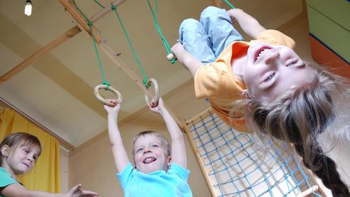 Drei Kinder spielen in einem Raum: Ein Junge hängt an Ringen, während zwei andere lachen und zuschauen.