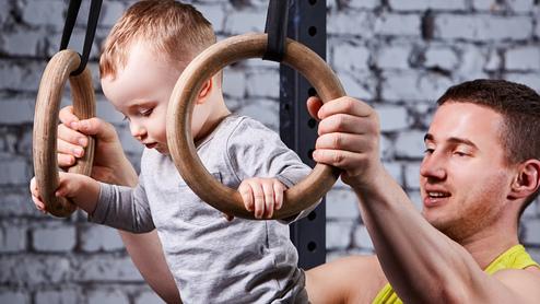 Ein Baby schwingt an Holzturnringen, während ein Erwachsener unterstützend daneben steht. Hintergrund mit Ziegelmauer.