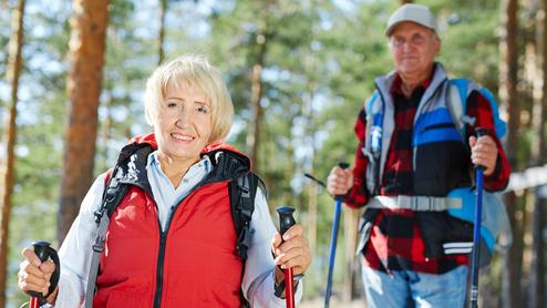 Zwei Wanderer in der Natur, eine Frau mit roter Weste und Wanderstock, ein Mann im Hintergrund mit Rucksack und Trekkingstöcken.