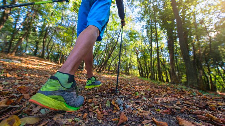 Nahaufnahme eines wandernden Mannes mit Wanderstöcken auf einem mit herbstlichen Blättern bedeckten Waldweg.