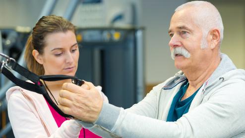 Ein älterer Mann mit Bart führt eine Übung mit einem Fitnessgerät unter Anleitung einer jungen Frau durch.