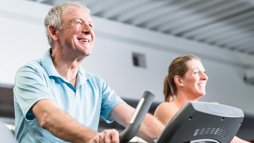 Älterer Mann und eine Frau trainieren lächelnd auf Ellipsentrainern in einem Fitnessstudio.