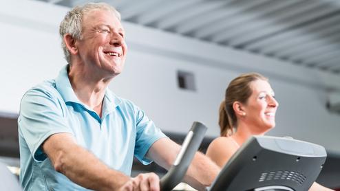 Älterer Mann und eine Frau trainieren lächelnd auf Ellipsentrainern in einem Fitnessstudio.