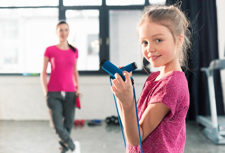Mädchen mit Sprungseil in der Hand, lächelt in einem Fitnessstudio, während eine Trainerin im Hintergrund steht.