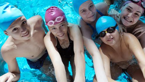 Gruppennahaufnahme von sieben Schwimmern im Wasser, lächelnd, mit Schwimmkappen und Brillen, vor einem blauen Hintergrund.