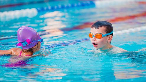 Zwei Kinder schwimmen in einem Schwimmbecken, eines tragt eine lilafarbene Badekappe und das andere orangefarbene Schwimmbrille.