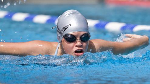 Junge Schwimmerin mit silberner Badekappe und Schwimmbrille in einem Wettkampfbecken, während sie einen Schlag ausführt.