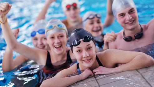Sechs Jugendliche in Badekappen und Schwimmbrillen posieren fröhlich im Wasser, einige mit erhobenen Armen.