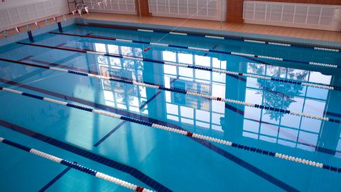Blick auf ein Schwimmbecken mit blauen Bahnen und reflektierendem Licht von Fenstern im Hintergrund.
