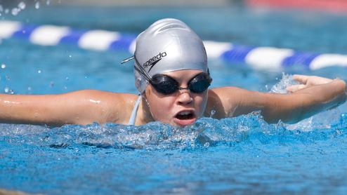 Junge Schwimmerin mit silberner Badekappe und Schwimmbrille in einem Wettkampfbecken, während sie einen Schlag ausführt.