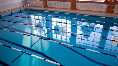 Blick auf ein Schwimmbecken mit blauen Bahnen und reflektierendem Licht von Fenstern im Hintergrund.
