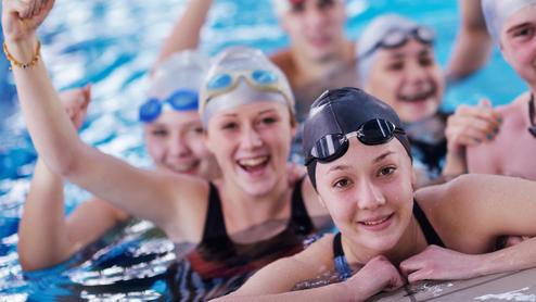 Sechs Jugendliche in Badekappen und Schwimmbrillen posieren fröhlich im Wasser, einige mit erhobenen Armen.